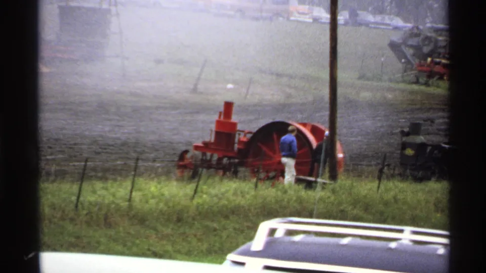 1971 Rollag, Minnesota frame showing a man near a red steam machine, fence posts, smoke haze, and parked vehicles.