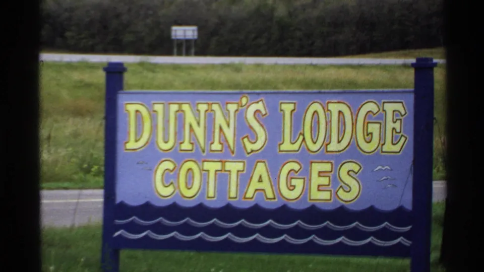 Close view of the Dunn's Lodge Cottages sign along a rural roadside, with grass and a road visible behind it.