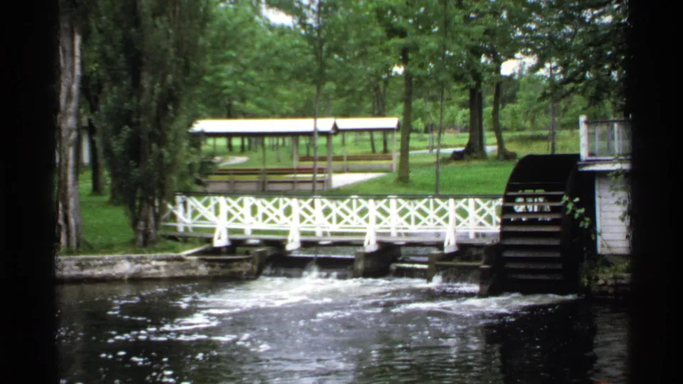 Riverside view at Dunn's Lodge Cottages with a white lattice bridge, wooden waterwheel, trees, lawn, and picnic shelter.