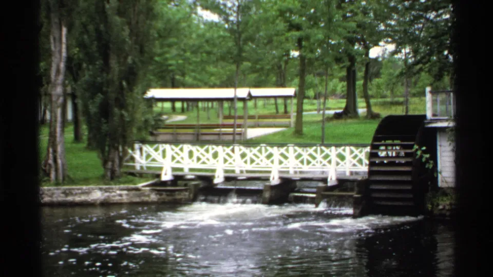 1971 home-movie view of the Rollag riverside picnic area with white bridge, waterwheel, trees, and covered shelter.