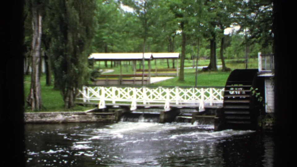 Riverside lodge grounds in Rollag, Minnesota, showing the white bridge, active water, wooden waterwheel, trees, and shelter.