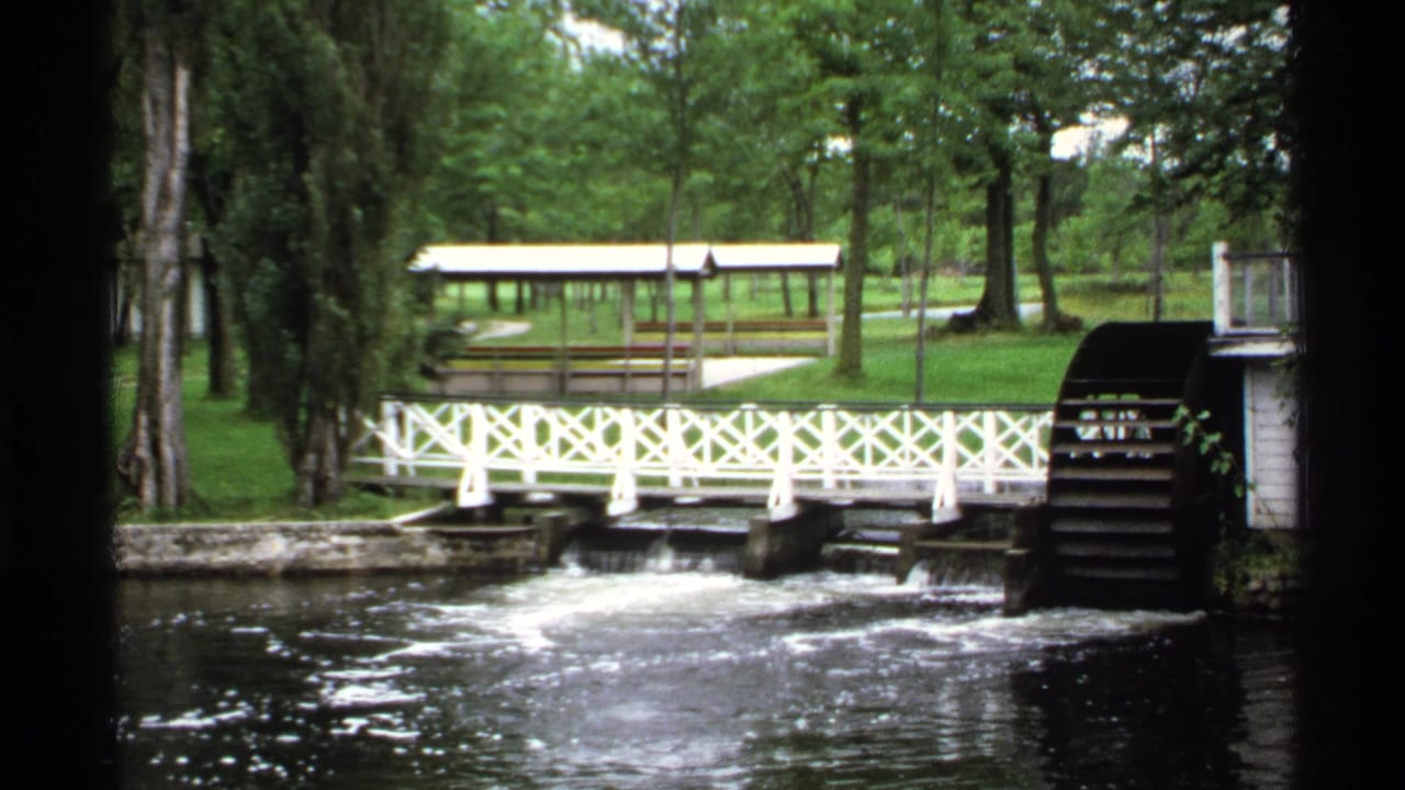 White pedestrian bridge over a stream beside a wooden waterwheel in a green park.