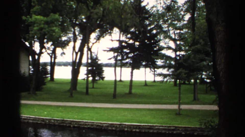 1971 Rollag, Minnesota 8mm frame showing a quiet lakeside lawn, tall trees, and a paved path with bright water beyond.