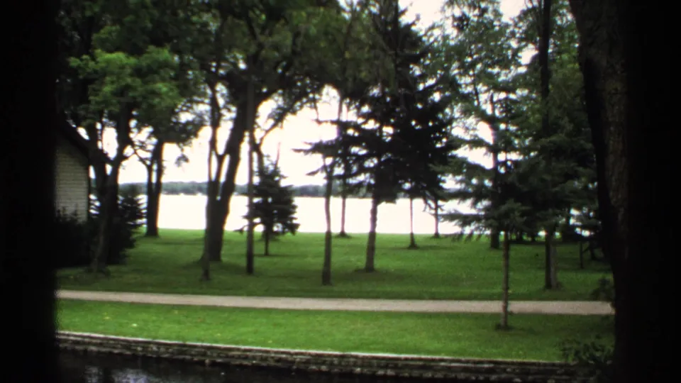 1971 Rollag, Minnesota 8mm frame showing a maintained lawn, paved path, and lakeside view through tall trees.