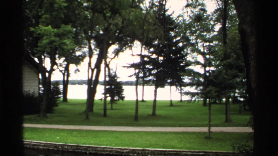 1971 Rollag, Minnesota 8mm frame of a lakeside park, with tall trees, a paved path, green lawn, and bright water in the distance.