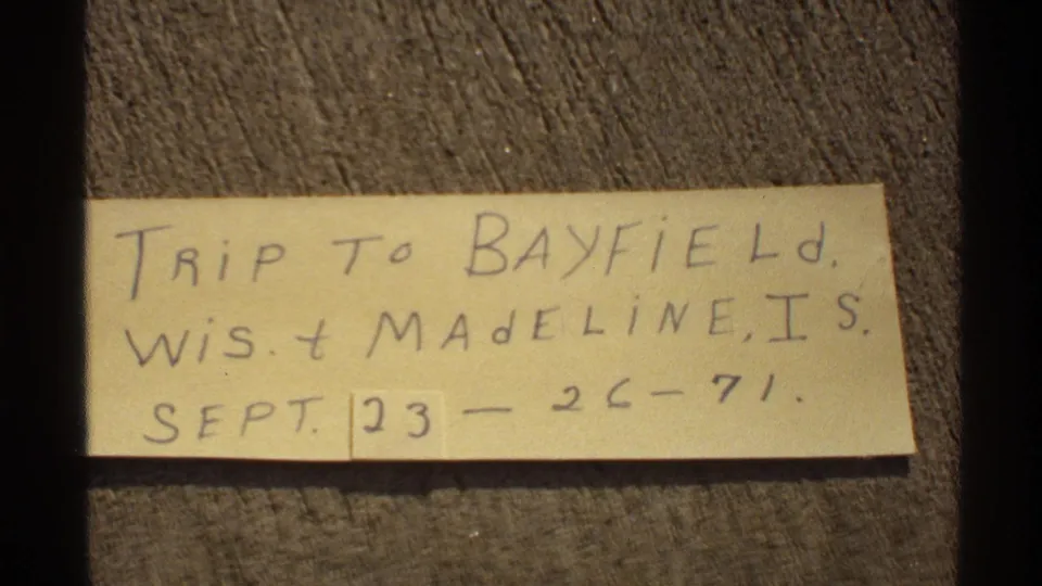 Handwritten 1971 trip card reading as a Bayfield, Wisconsin and Madeline Island note, filmed on a wood surface.