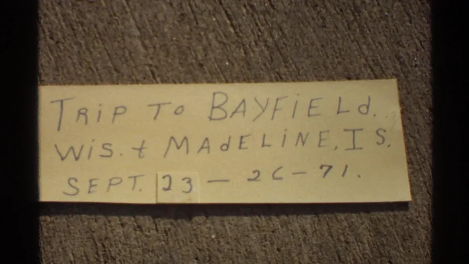 1971 home-movie frame of a handwritten trip label for Bayfield, Wisconsin and Madeline Island on a wood surface.