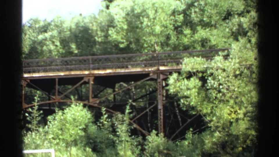 A weathered metal truss bridge spans a wooded area in this 1971 Minnesota-linked 8mm home-movie frame.