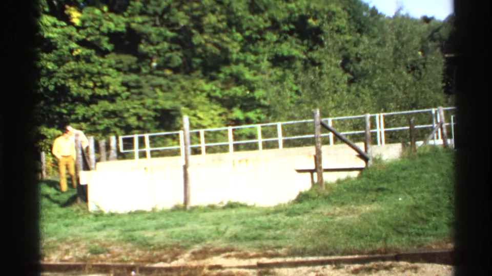 Two distant people stand near a low concrete structure and metal railing in a grassy rural area in Minnesota-linked 1971 footage.