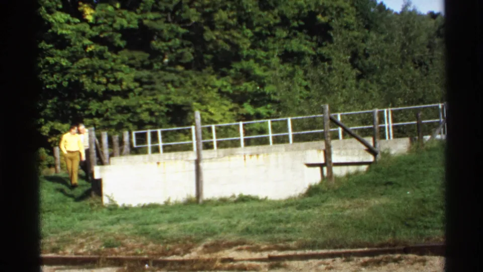 1971 Rollag, Minnesota home-movie frame showing two people near a concrete retaining wall, railing, grass, and trees.