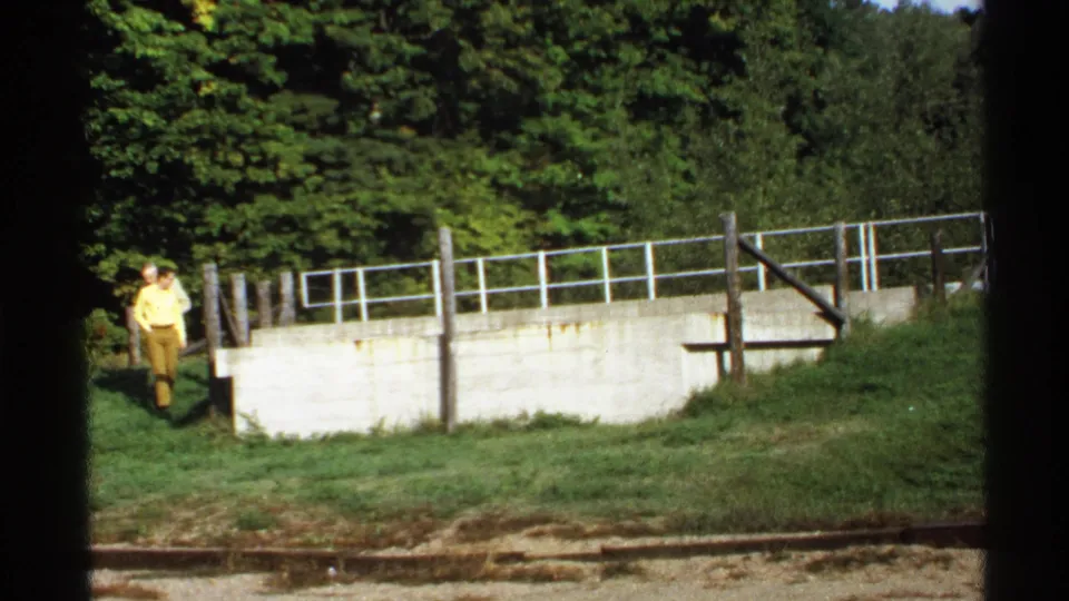 1971 Rollag, Minnesota frame with a person in a yellow shirt by a concrete wall, white railing, grass, and dense trees.