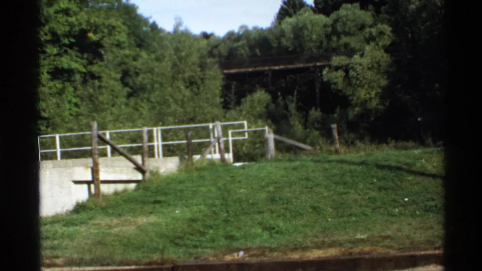 1971 Rollag, Minnesota home-movie view of a grassy slope, concrete wall, white railing, and wooded backdrop.
