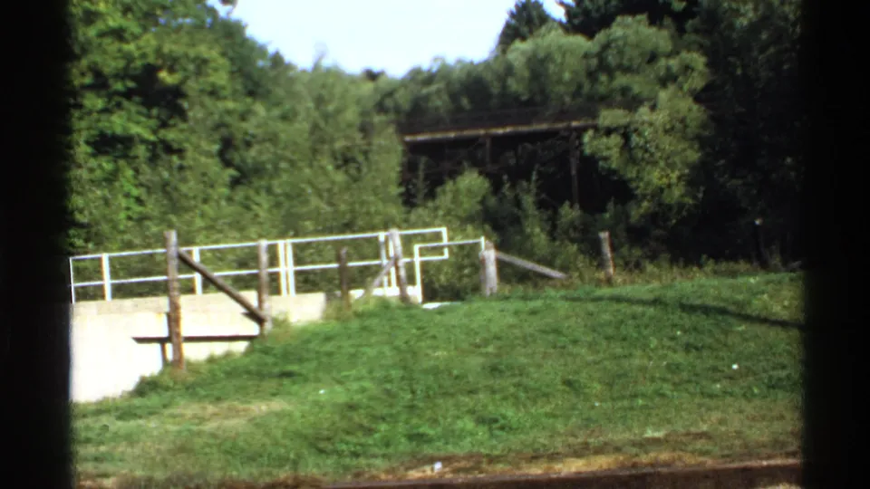 1971 Rollag, Minnesota frame showing grass, a concrete retaining wall, white railing, wooden posts, and a wooded background.