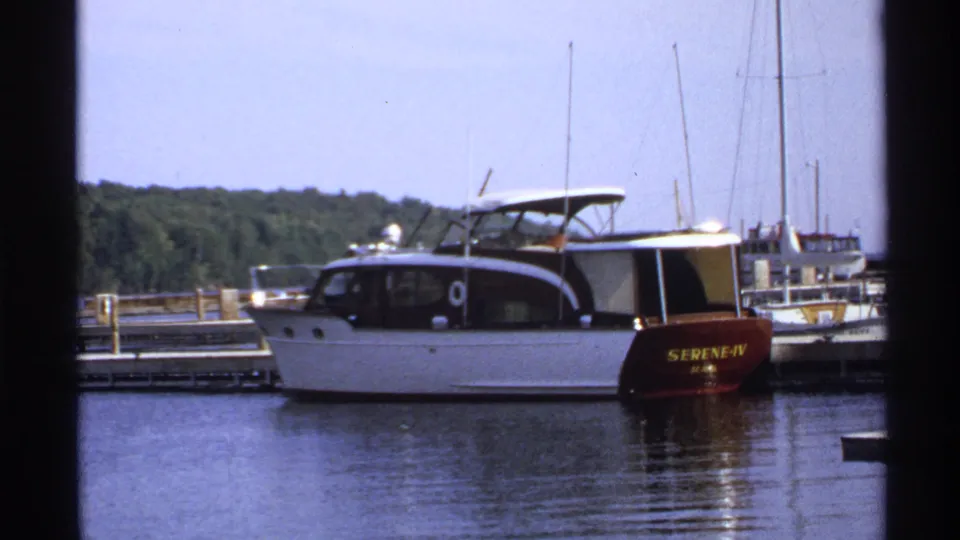 1971 Rollag, Minnesota 8mm frame of a white motorboat docked beside piers, with sailboat masts and a wooded shoreline in view.