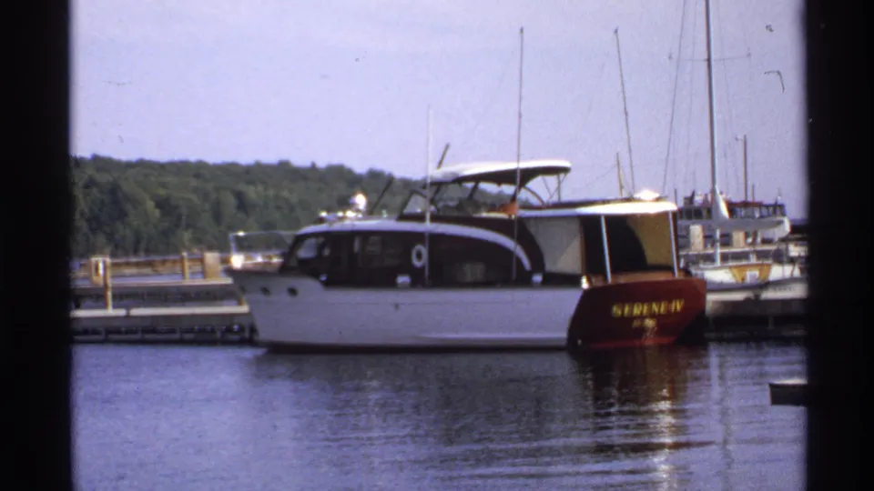 1971 Rollag, Minnesota 8mm frame of the docked SERENE-IV motorboat on calm water with marina masts and trees behind it.