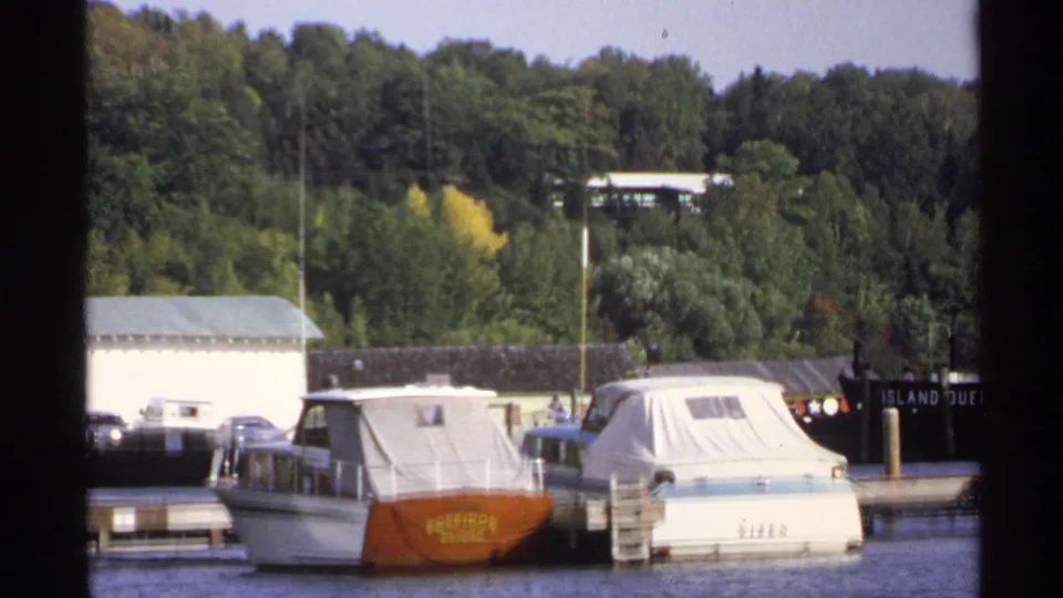 1971 Rollag, Minnesota harbor view with covered cabin boats, dock pilings, wooded shoreline, and a dark boat bearing visible lettering.