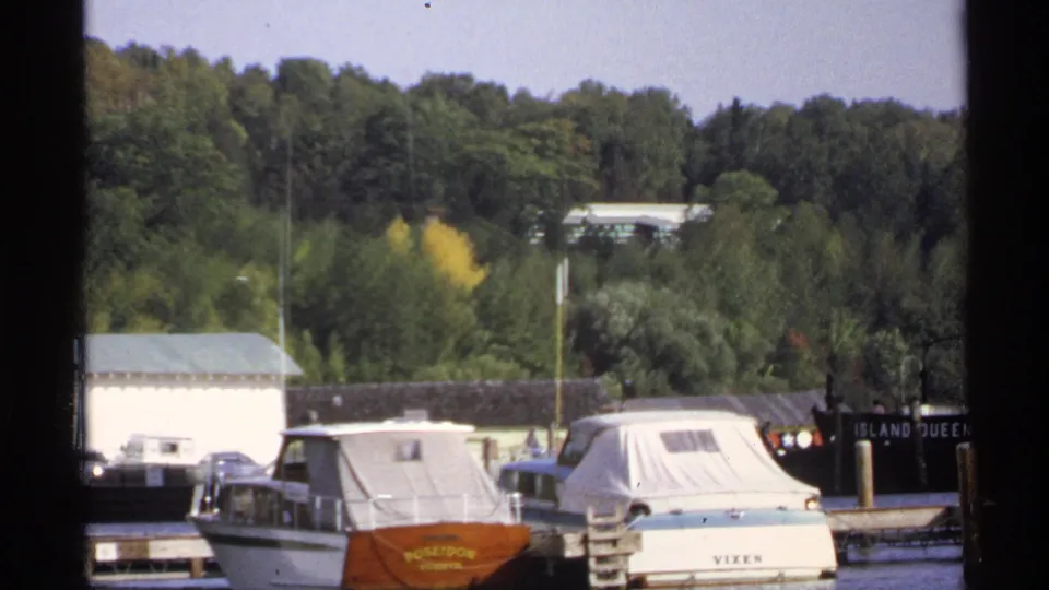 1971 Rollag, Minnesota 8mm harbor frame with covered boats, a white dockside building, dense trees, and readable vessel markings.