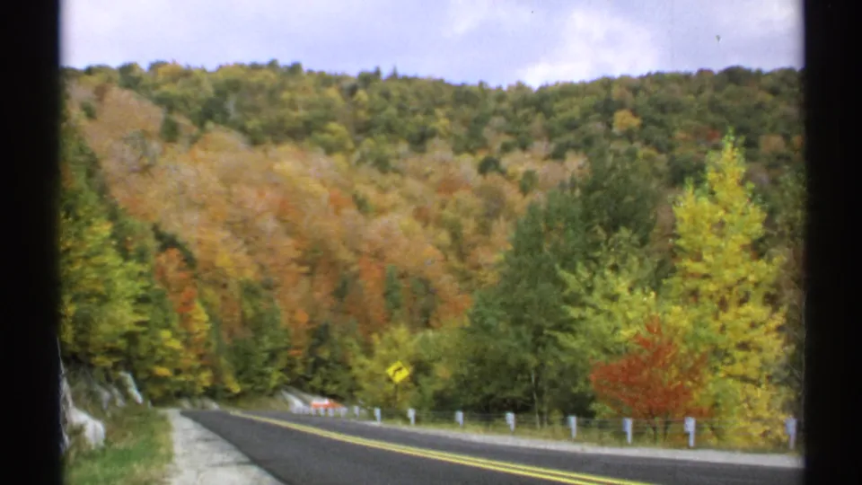 1971 Vermont roadside view of a paved curve, fall-colored woods, and a small car partly visible near the bend.