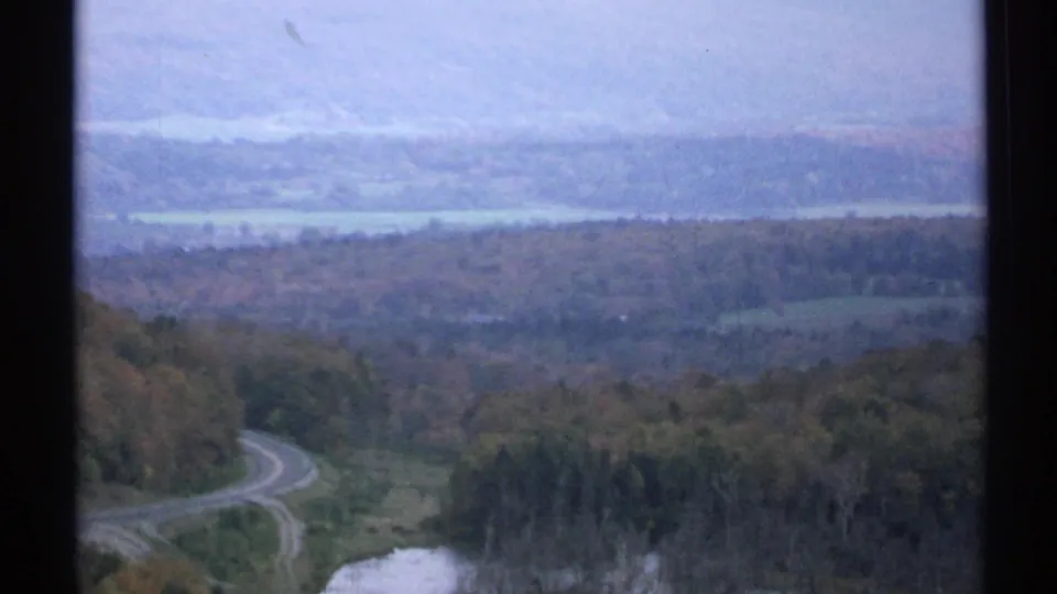 1971 Vermont 8mm wide view of wooded hills, a curving rural road, and a pale distant water or field strip.