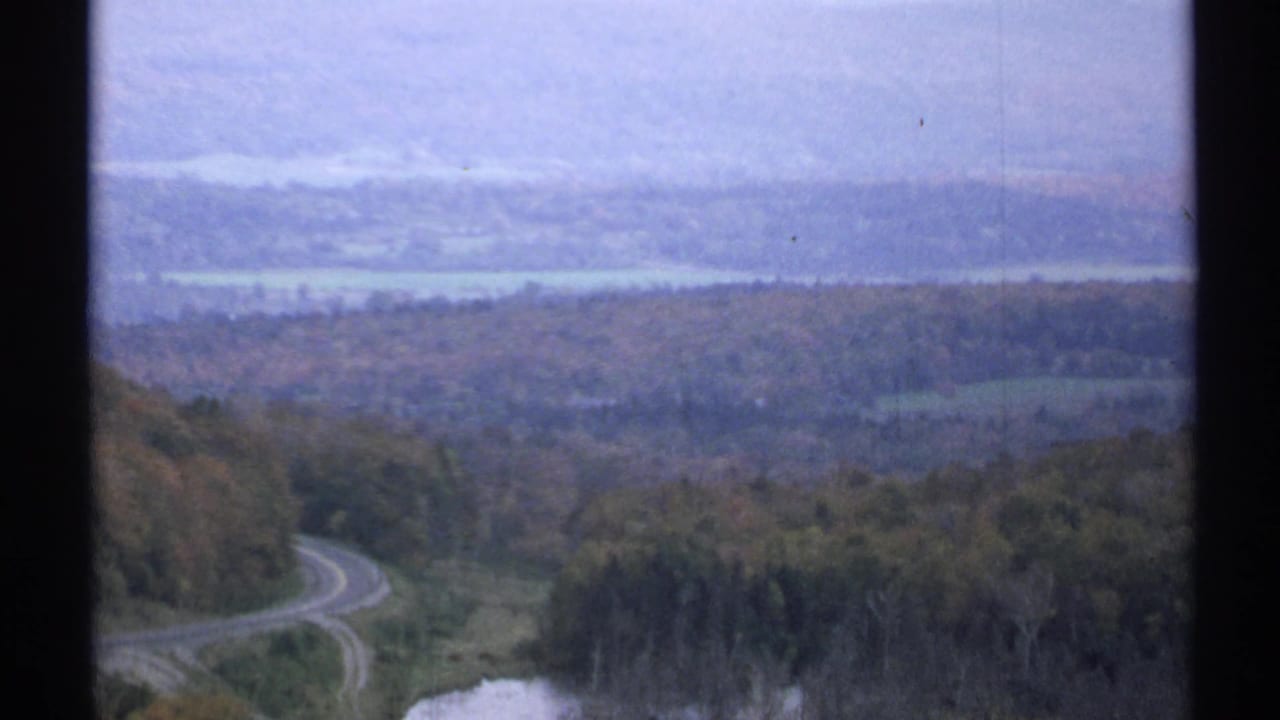 Winding rural road through autumn woods with distant water and hazy hills.