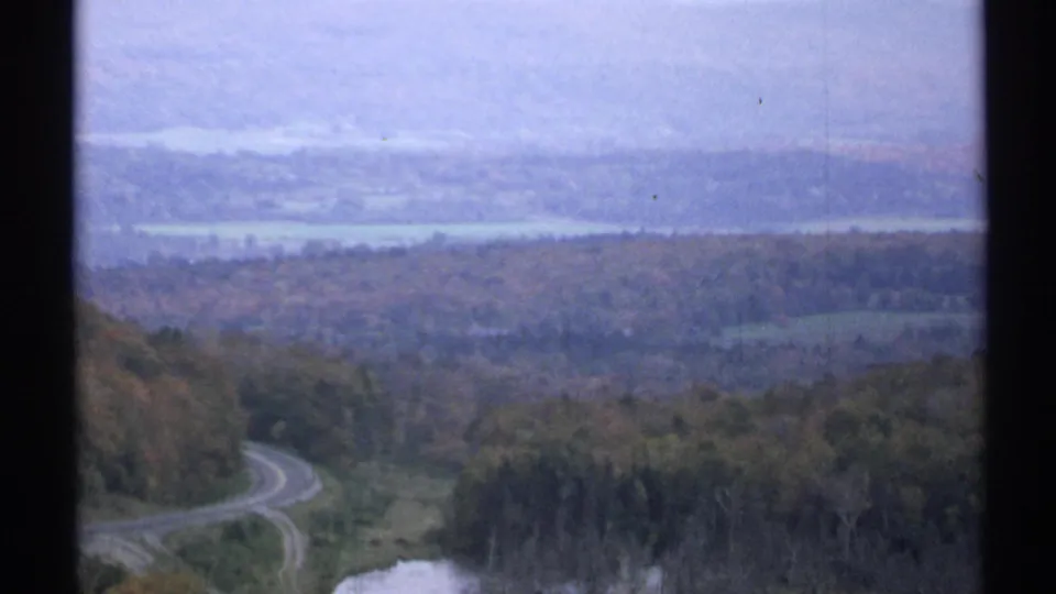 1971 Vermont 8mm frame showing a curving country road, fall-colored forest, distant water, and hazy hills beyond a dark film border.