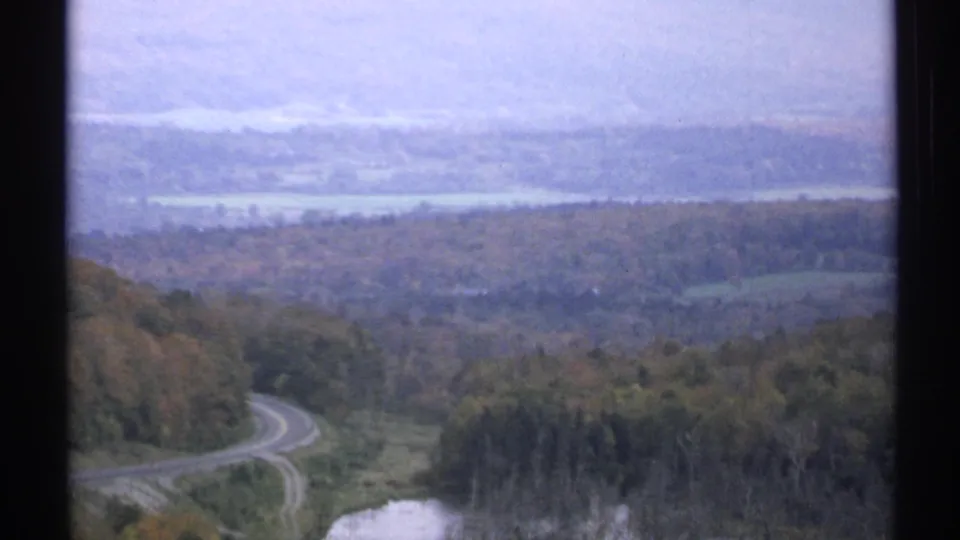 1971 Vermont home-movie view from above a wooded valley, with a curving road below and pale water visible under a hazy sky.
