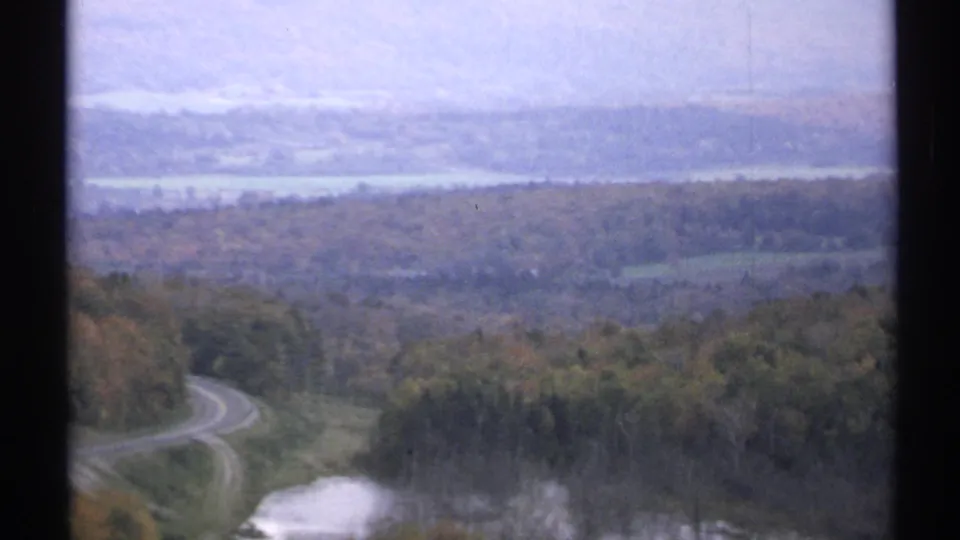 Soft 1971 Vermont 8mm frame with a road bend at lower left, dense fall woods, reflective water below, and distant hills.