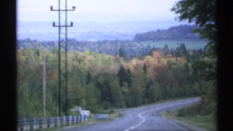 1971 Vermont 8mm frame from a car view down a winding rural road with utility poles, guardrails, trees, hills, and pale distant mountains.