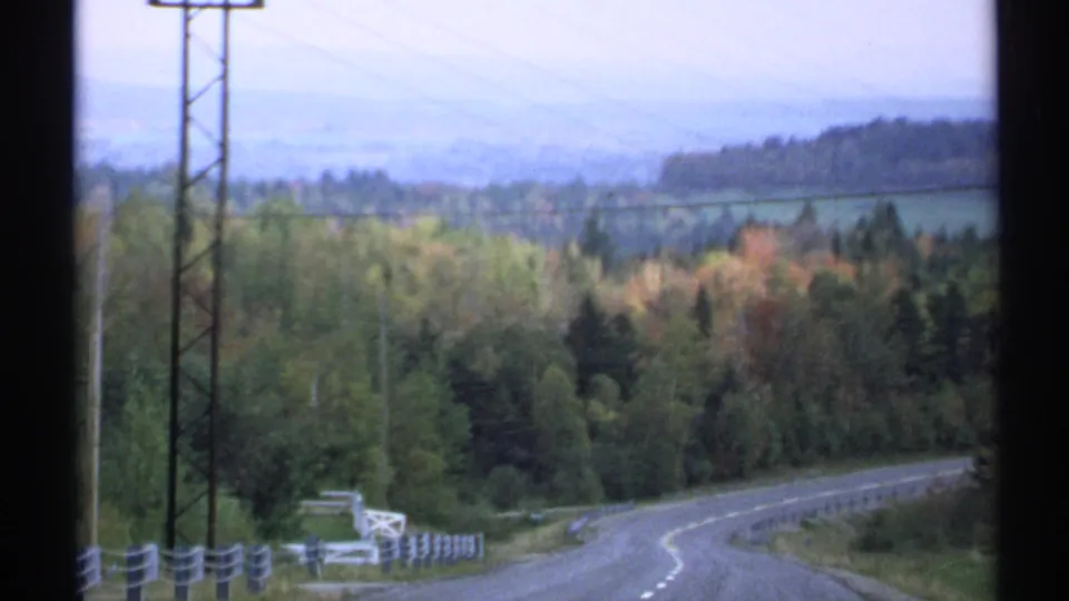 1971 Vermont 8mm frame looking down a quiet curving road bordered by guardrails, utility poles, mixed trees, hills, and distant mountains.