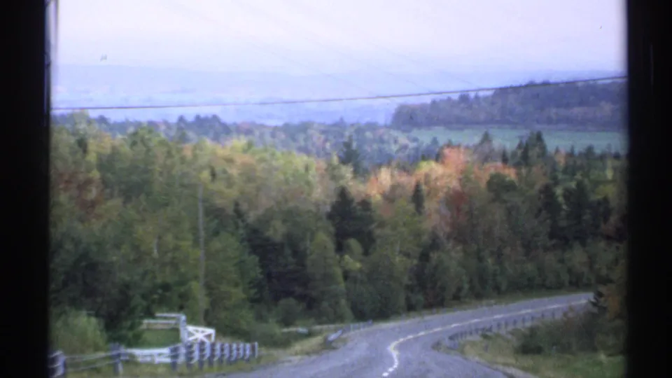 1971 Vermont 8mm view of a winding rural road with autumn trees, roadside guardrail, utility lines, and hazy hills beyond.