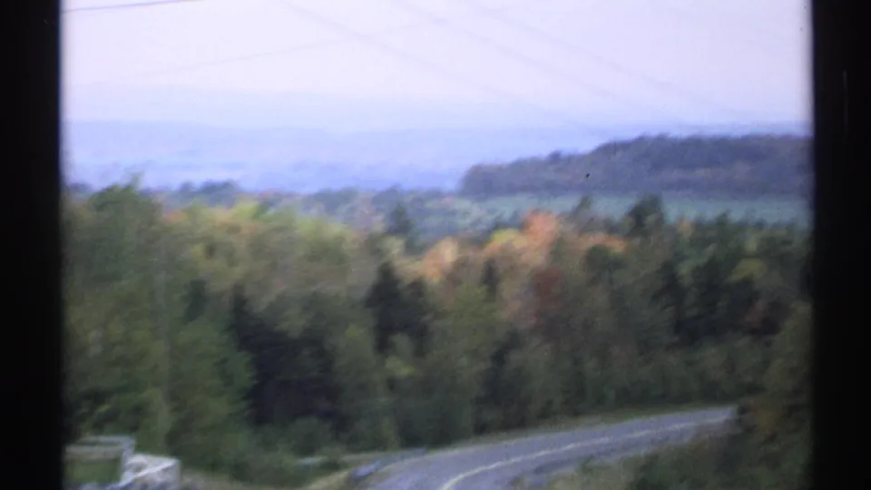 Soft 8mm frame from a Vermont drive showing a road bend, roadside trees, utility wires, and wooded hills in the distance.