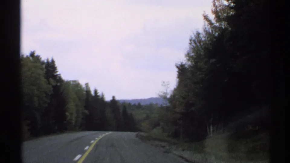 1971 Vermont driving view along a two-lane road bordered by evergreens, leafy trees, distant low hills, and an overcast sky.