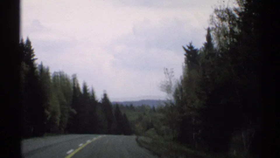 1971 Vermont road view from a moving car, with pine woods, a centerline, and distant low hills.