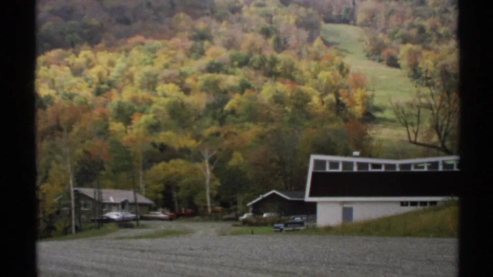 1971 Vermont 8mm frame looking from the resort area toward wooded hills, gravel foreground, buildings, and parked cars.