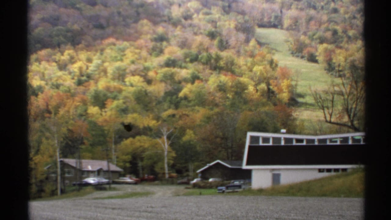 Gravel lot with buildings below a wooded hillside in fall color.