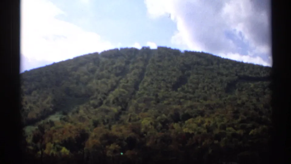 1971 Vermont 8mm hillside view with dense green forest, autumn color near the base, and a bright cloudy sky.