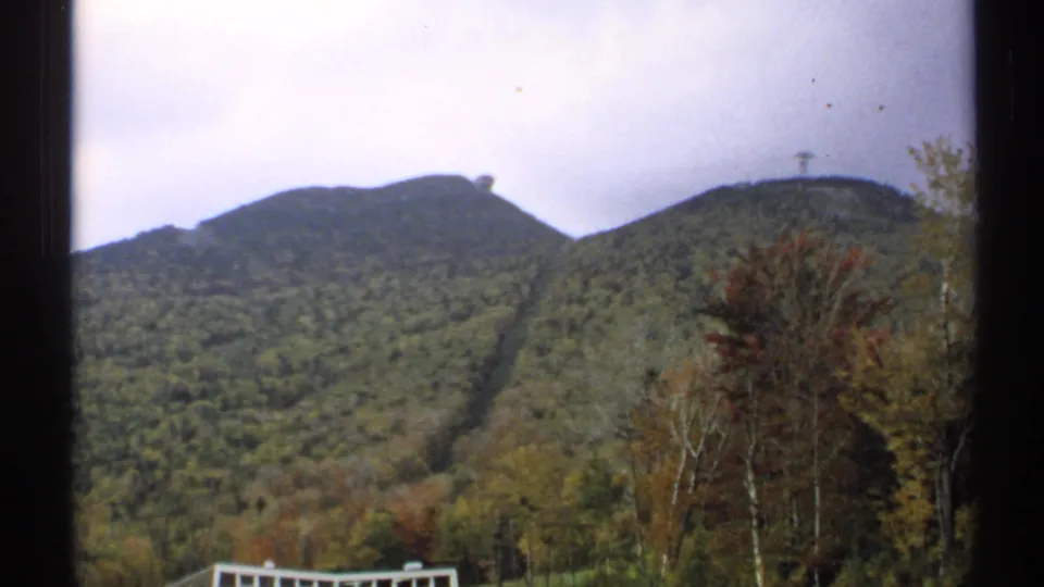 1971 Vermont 8mm frame showing rounded mountain peaks, a ski-lift clearing, autumn trees, a pale foreground railing, and cloudy sky.