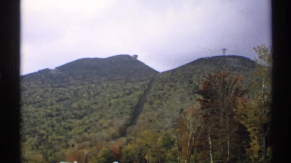 1971 Vermont 8mm view of forested mountain slopes, fall foliage, a ski-lift corridor, small summit structures, and dark film framing.