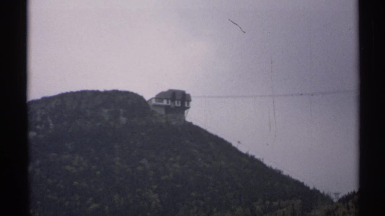 Forested Vermont hill with a small building near the ridge under a pale cloudy sky.
