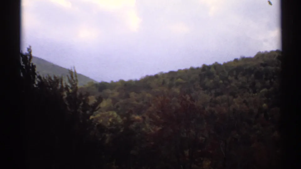 1971 Vermont 8mm landscape frame with dense woodland, a sloping mountain form, and pale clouds overhead.