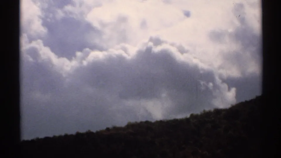 1971 Vermont 8mm cloudscape with bright puffy clouds over a shadowed wooded hillside.