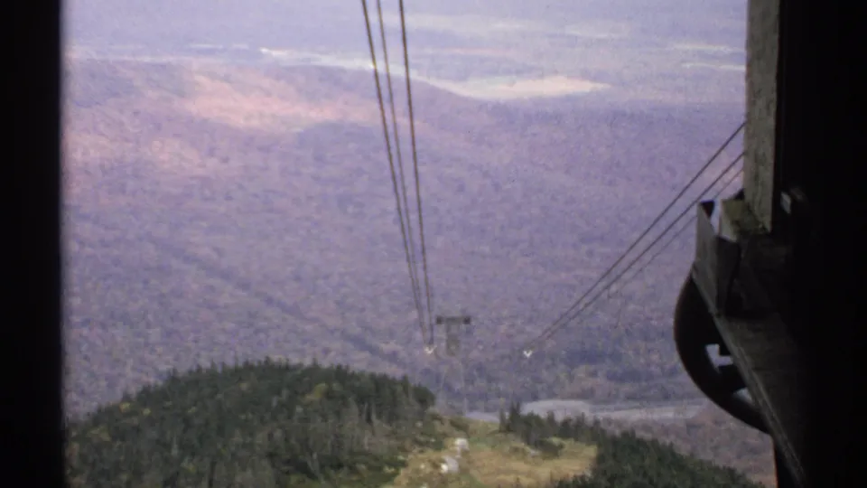 1971 Vermont home-movie frame with cable lines, a support tower, tram hardware, and a hazy forested mountain valley.