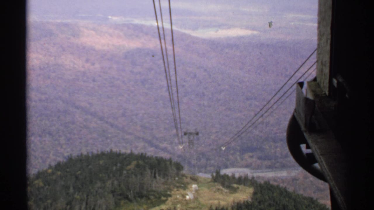 1971 Vermont 8mm view from a tram area, with cable lines crossing a forested slope toward a distant mountain valley.