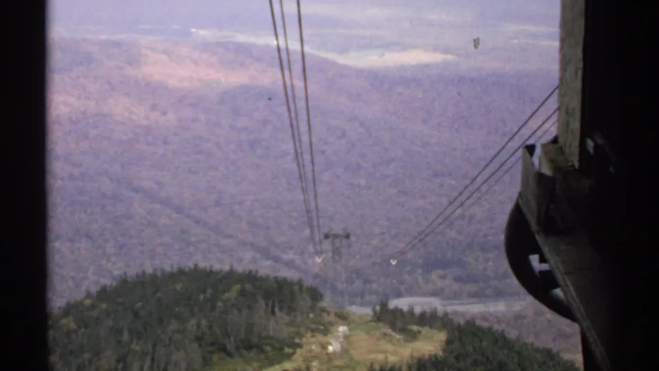1971 Vermont 8mm view from a tram area, with cable lines crossing a forested slope toward a distant mountain valley.