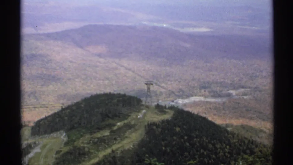 1971 Vermont 8mm frame looking from a mountaintop over forested hills, a wide valley, distant slopes, and a cable car tower.