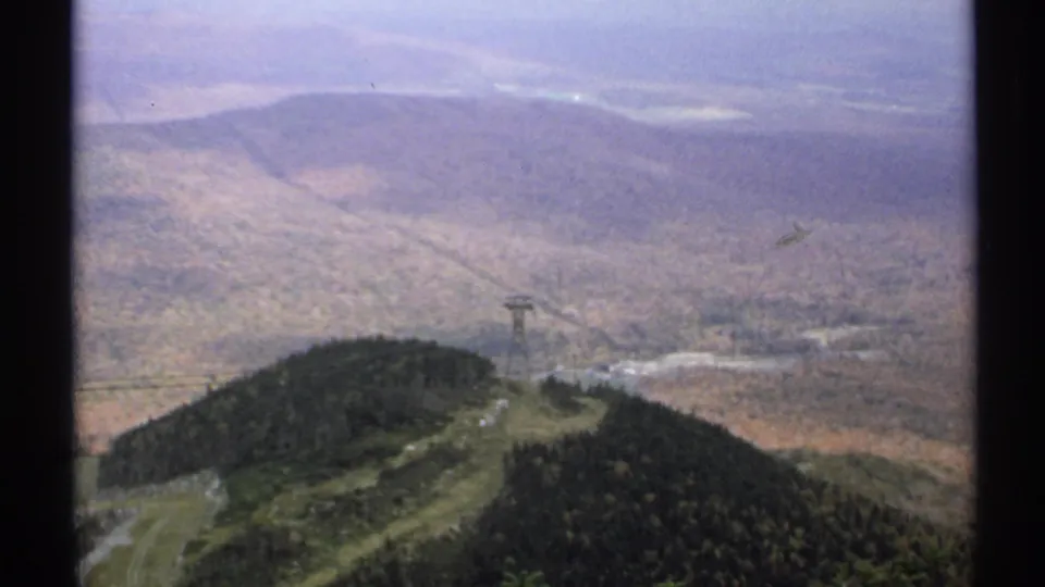 1971 Vermont frame showing a hazy mountain panorama with a wooded ridge, valley floor, distant slopes, and a visible cable tower.