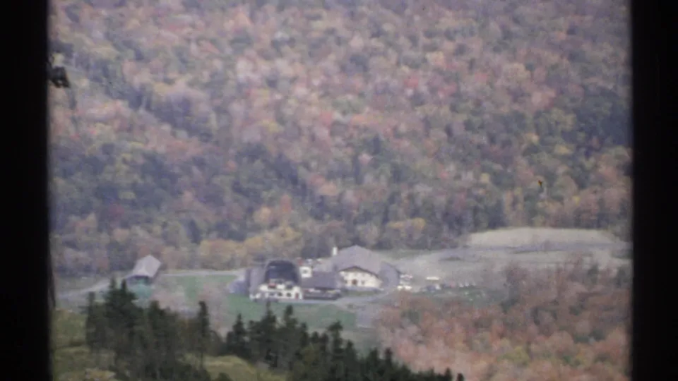Final 1971 Vermont frame showing resort-like valley buildings beneath a wooded fall hillside, with soft 8mm grain.