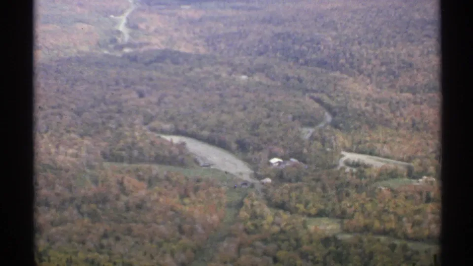Elevated 1971 Vermont view of autumn hills, a curved rural road, a water feature, and scattered buildings below.