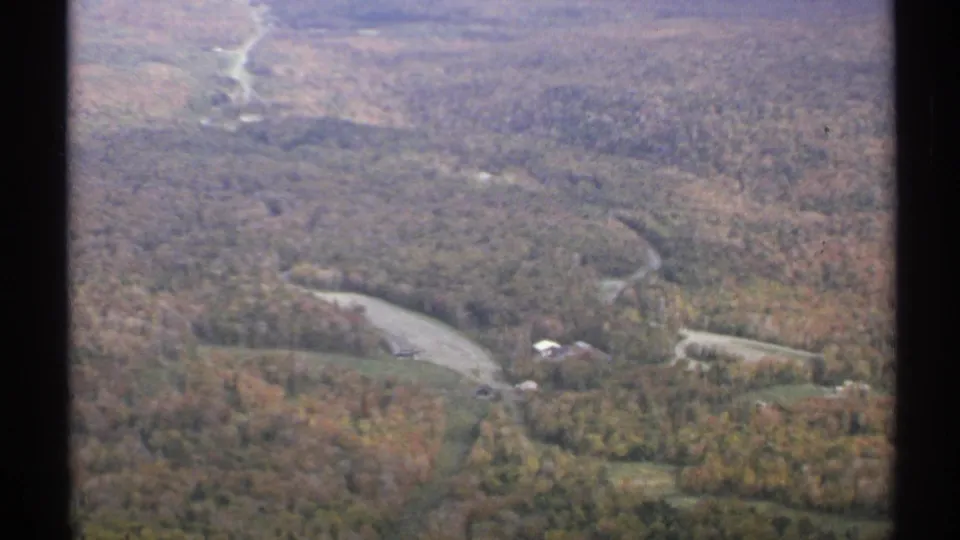 1971 Vermont aerial home-movie frame showing a wooded autumn valley, water bend, road, and distant hillside terrain.