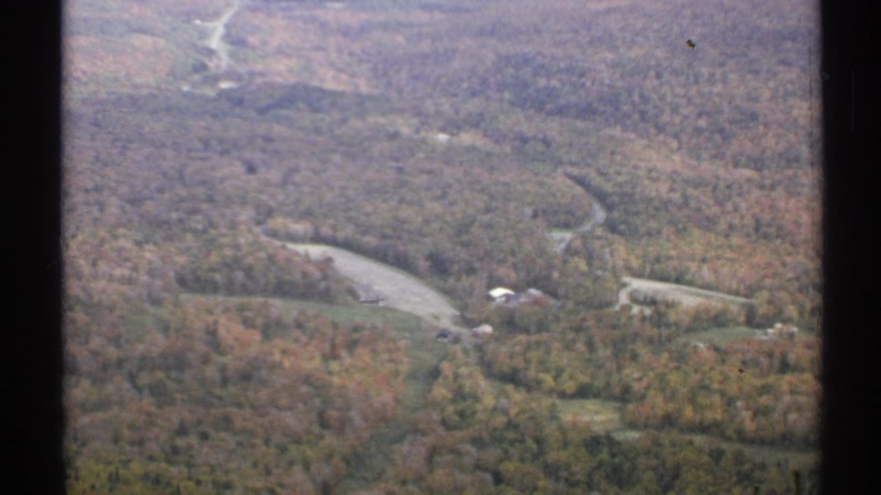 High view over a forested valley with a winding road, fields, and small farm buildings.